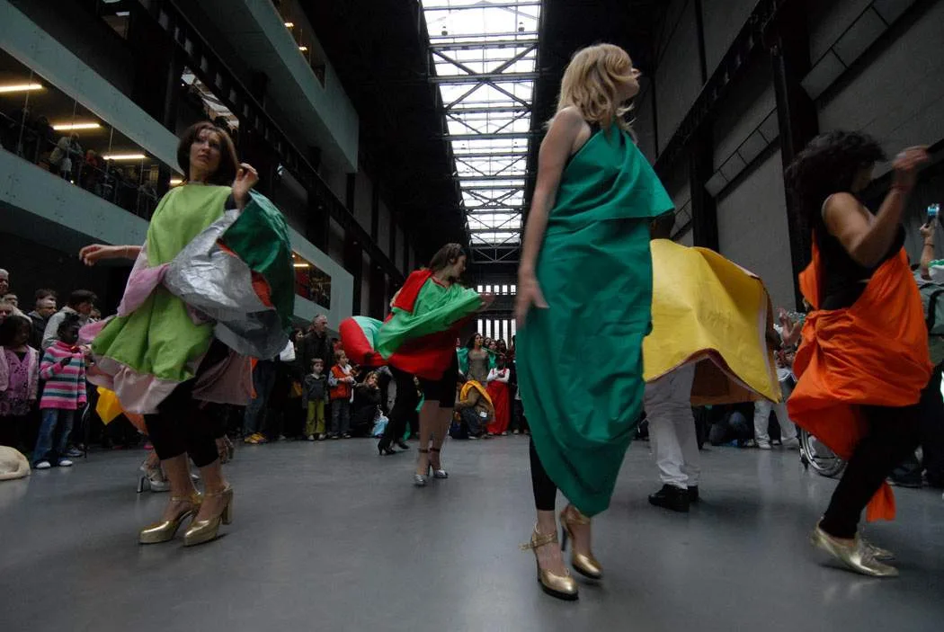 Dancers in green, yellow, orange and patterned fabric capes moving across the floor of the Turbine Hall at Tate Modern during a 2007 restaging of Hélio Oiticica's Parangolés